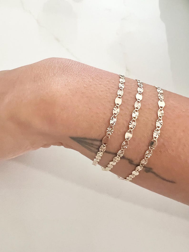May include: Three delicate gold bracelets, each featuring a repeating pattern of oval and star-shaped links. The bracelets are displayed on a person's arm, showcasing their intricate design and reflective surface. The jewelry is set against a neutral background.