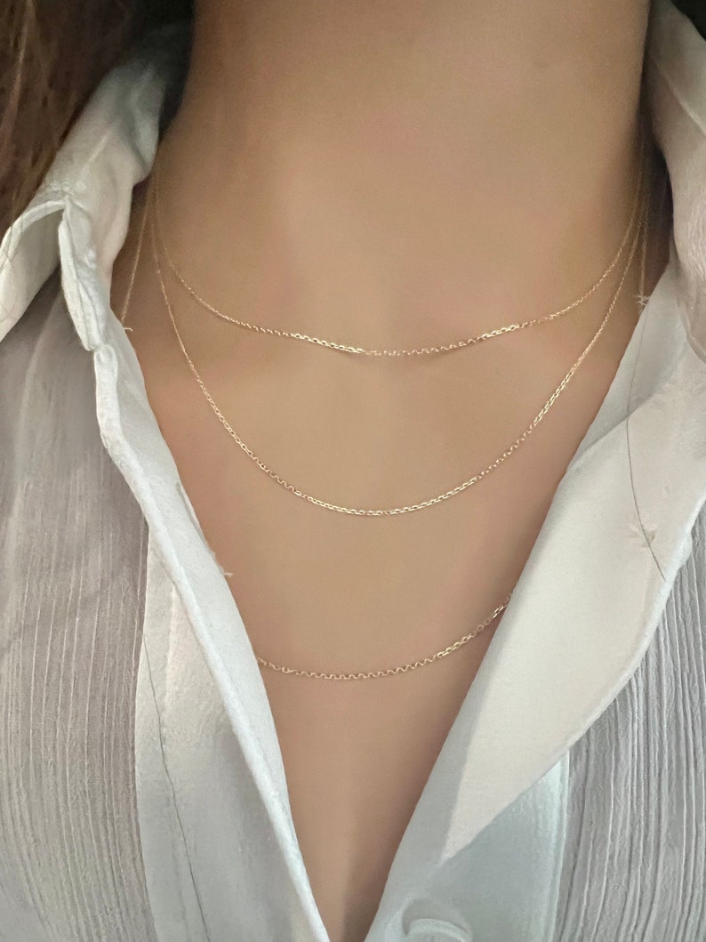 May include: Three gold chain necklaces layered on a person's neck. The chains are delicate and have a simple design.