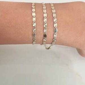May include: Three delicate gold bracelets, each featuring a repeating pattern of oval and star-shaped links. The bracelets are displayed on a person's wrist, showcasing their intricate design and warm, golden hue. The bracelets are a classic jewelry item.