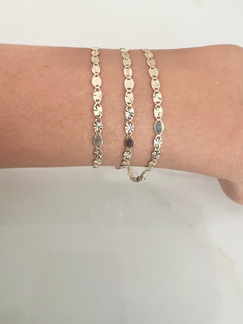 May include: Three delicate gold bracelets, each featuring a repeating pattern of oval and star-shaped links. The bracelets are displayed on a person's wrist, showcasing their intricate design and warm, golden hue. The bracelets are a classic jewelry item.
