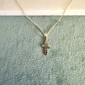May include: A gold-colored pendant necklace featuring a small, ornate cross. The cross has black detailing and is suspended from a delicate chain. The background is a soft, textured teal color.