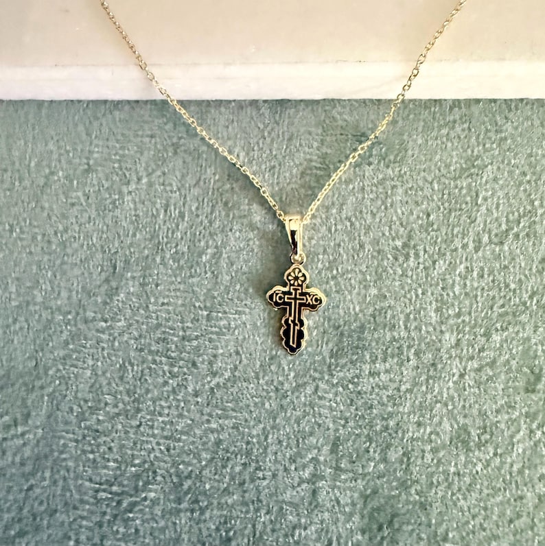 May include: A gold-colored pendant necklace featuring a small, ornate cross. The cross has black detailing and is suspended from a delicate chain. The background is a soft, textured teal color.