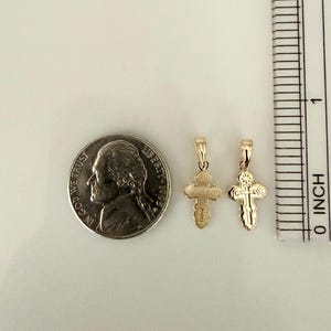 May include: Two small gold-colored cross pendants with a coin and ruler for scale. The crosses have intricate designs and are suspended from small loops. The ruler shows the crosses are less than 1 inch in length.