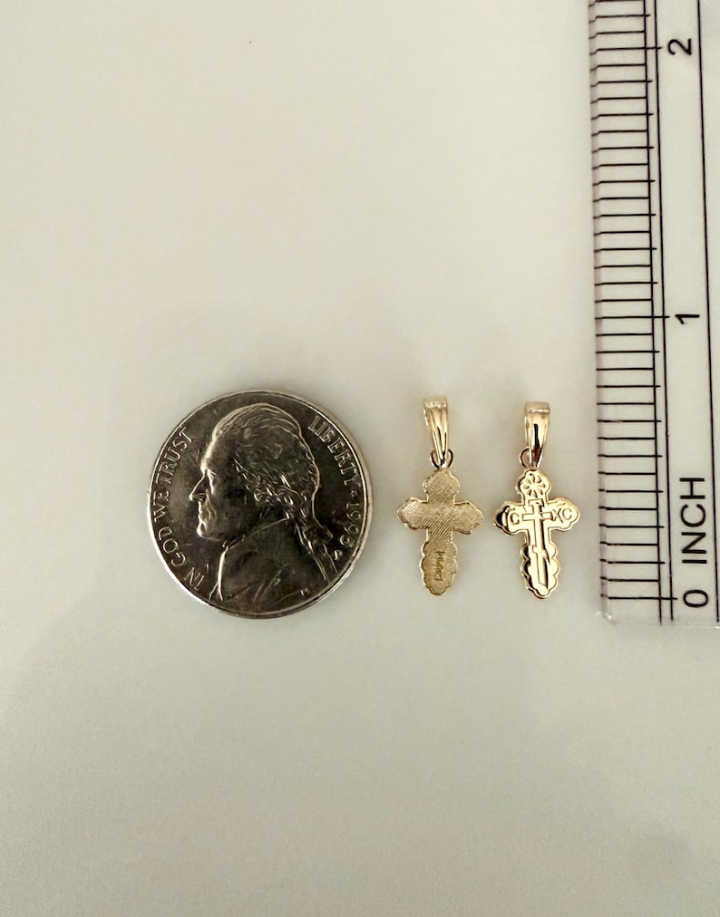 May include: Two small gold-colored cross pendants with a coin and ruler for scale. The crosses have intricate designs and are suspended from small loops. The ruler shows the crosses are less than 1 inch in length.