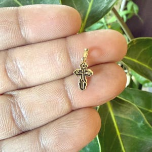 May include: A small, gold-colored pendant in the shape of a cross. The cross has intricate black detailing and a loop at the top for a chain. The pendant is held in a hand against a background of green leaves.