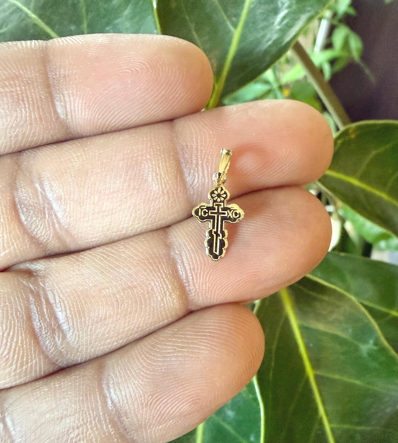 May include: A small, gold-colored pendant in the shape of a cross. The cross has intricate black detailing and a loop at the top for a chain. The pendant is held in a hand against a background of green leaves.