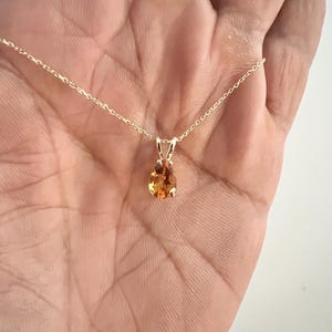 May include: A gold necklace with a small, oval-shaped, orange gemstone pendant. The pendant is suspended from a delicate gold chain. The gemstone has a warm, amber color and is set in a simple gold setting.