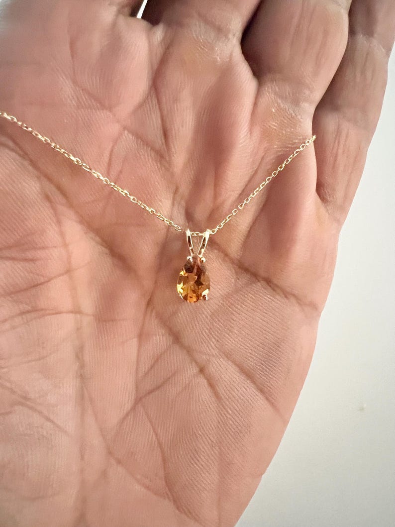 May include: A gold necklace with a small, oval-shaped, orange gemstone pendant. The pendant is suspended from a delicate gold chain. The gemstone has a warm, amber color and is set in a simple gold setting.