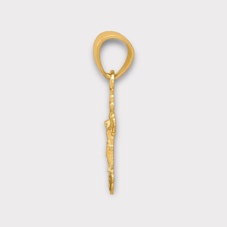 May include: A gold-coloured pendant depicting a person on a cross. The pendant has a loop at the top for a chain. The background is a light grey colour.
