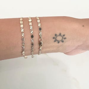 May include: Three delicate gold bracelets with various linked designs, including circles, ovals, and floral patterns. The bracelets are displayed on a light-toned background. A small sun-shaped tattoo is visible on the wrist.