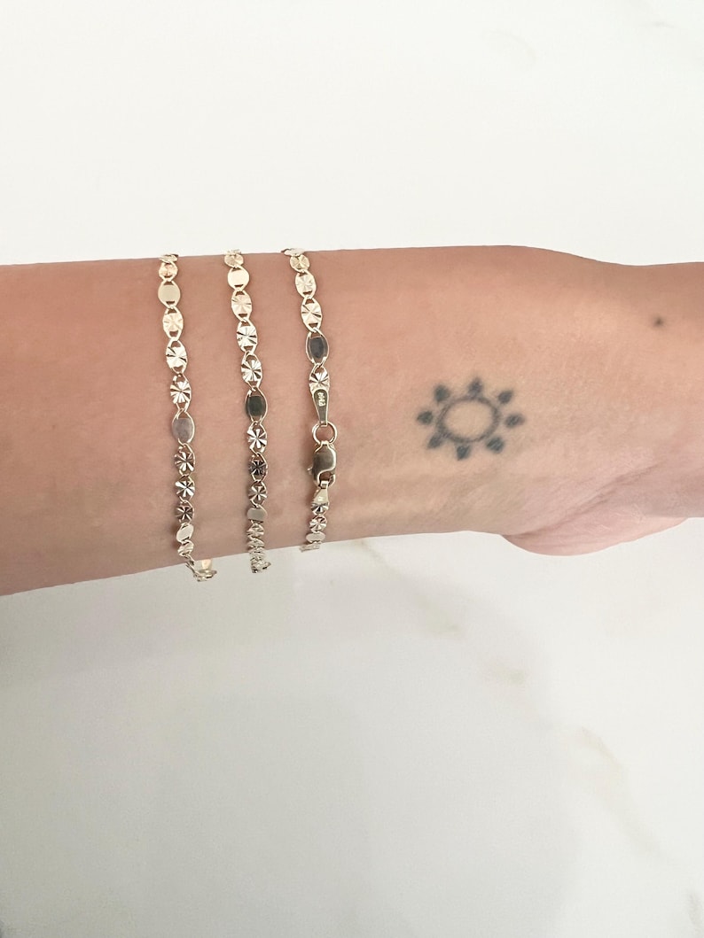 May include: Three delicate gold bracelets with various linked designs, including circles, ovals, and floral patterns. The bracelets are displayed on a light-toned background. A small sun-shaped tattoo is visible on the wrist.