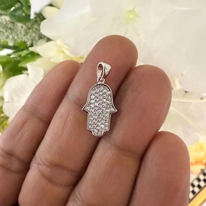 May include: A silver-colored Hamsa hand pendant, encrusted with small, sparkling clear stones. The pendant is held against a background of white flowers. The Hamsa hand is a symbol of protection and good fortune.
