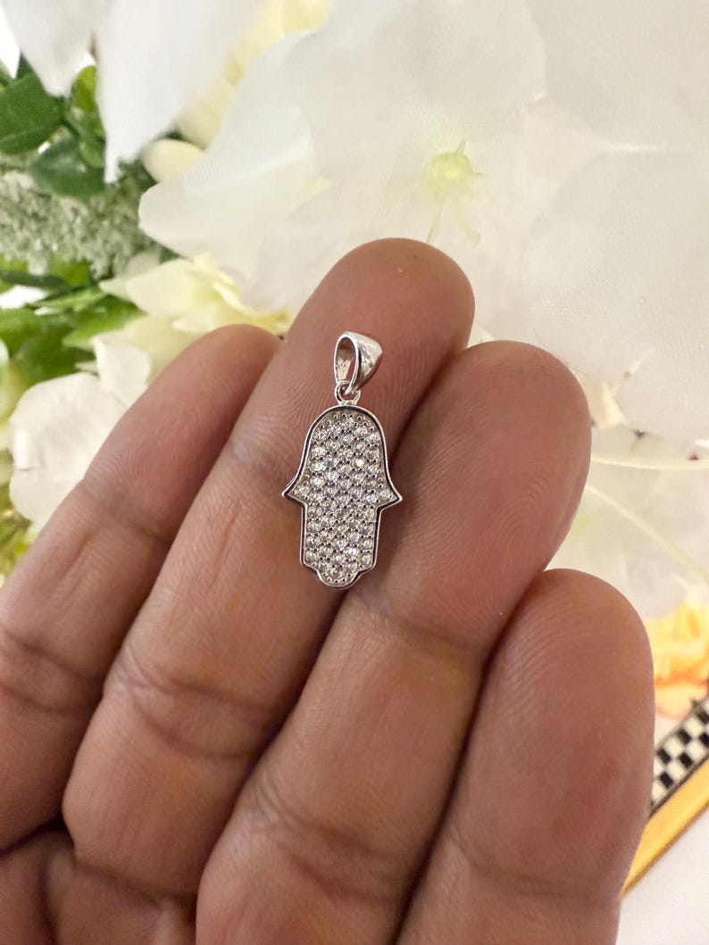 May include: A silver-colored Hamsa hand pendant, encrusted with small, sparkling clear stones. The pendant is held against a background of white flowers. The Hamsa hand is a symbol of protection and good fortune.