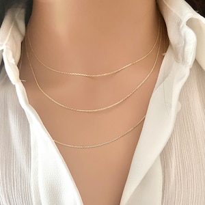 May include: Three gold chain necklaces layered on a model's neck. The chains are delicate and have a simple design.