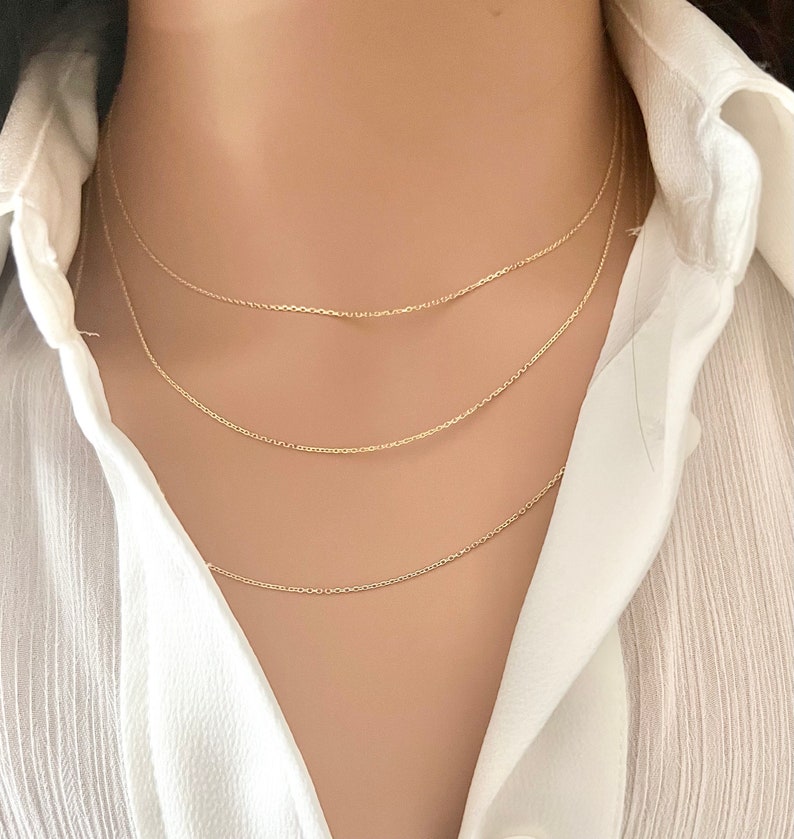 May include: Three gold chain necklaces layered on a model's neck. The chains are delicate and have a simple design.