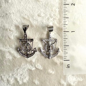 K&ouml;nnte beinhalten: Zwei silberfarbene Ankeranh&auml;nger mit klaren Edelsteinakzenten. Die Anh&auml;nger haben ein nautisches Design mit einem Kreuz und einem Steuerrad. Sie werden auf einer wei&szlig;en, strukturierten Oberfl&auml;che neben einem Zoll-Lineal pr&auml;sentiert.