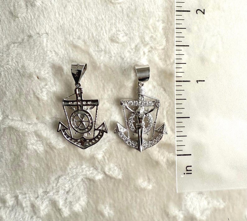 K&ouml;nnte beinhalten: Zwei silberfarbene Ankeranh&auml;nger mit klaren Edelsteinakzenten. Die Anh&auml;nger haben ein nautisches Design mit einem Kreuz und einem Steuerrad. Sie werden auf einer wei&szlig;en, strukturierten Oberfl&auml;che neben einem Zoll-Lineal pr&auml;sentiert.