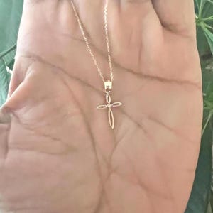 Puede incluir: Un delicado collar dorado con un colgante de cruz. La cruz tiene un dise&ntilde;o abierto y minimalista. El collar se muestra sobre un fondo de tono de piel claro, con un fondo verde borroso.