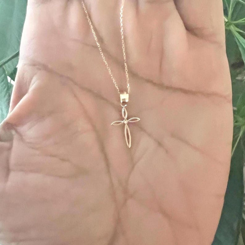 Puede incluir: Un delicado collar dorado con un colgante de cruz. La cruz tiene un dise&ntilde;o abierto y minimalista. El collar se muestra sobre un fondo de tono de piel claro, con un fondo verde borroso.