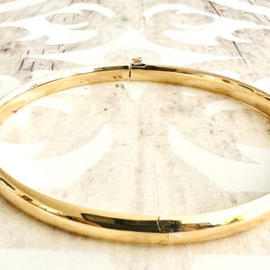 May include: A gold-colored bangle bracelet with a hinged clasp. The bracelet is circular and has a smooth, polished surface. The jewelry is displayed on a light-colored surface with a subtle pattern.