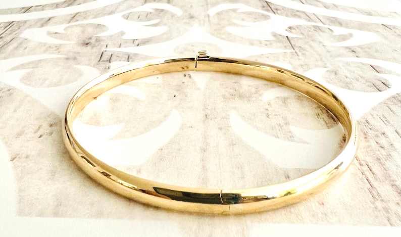May include: A gold-colored bangle bracelet with a hinged clasp. The bracelet is circular and has a smooth, polished surface. The jewelry is displayed on a light-colored surface with a subtle pattern.