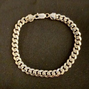 May include: A silver curb chain bracelet with a secure clasp. The bracelet features a series of interlocking, rounded links. The metal has a bright, reflective finish. The bracelet is displayed against a dark background.
