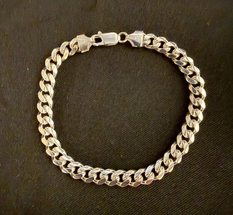 May include: A silver curb chain bracelet with a secure clasp. The bracelet features a series of interlocking, rounded links. The metal has a bright, reflective finish. The bracelet is displayed against a dark background.