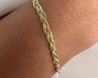 May include: A gold braided chain bracelet. The bracelet is made of a thick, braided chain that is about 1/2 inch wide.