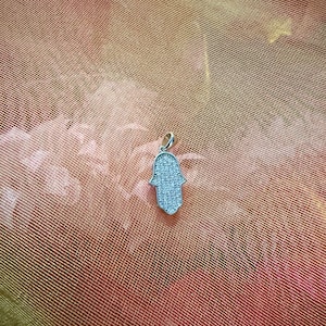 May include: A silver-colored Hamsa hand pendant, encrusted with small, sparkling stones. The pendant is displayed against a textured, peach-colored background, creating a contrast that highlights the jewelry's details.