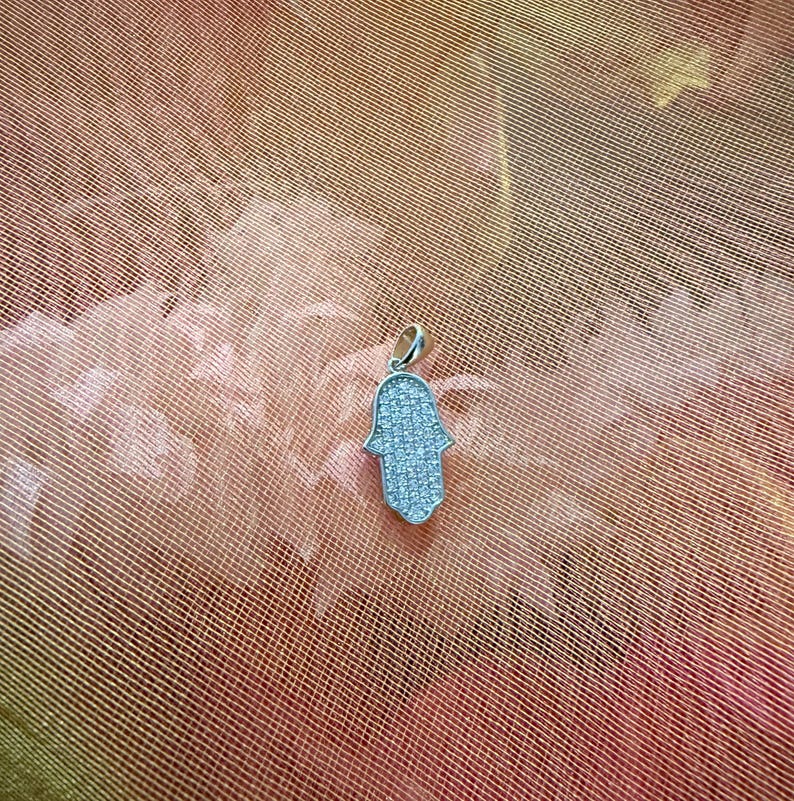 May include: A silver-colored Hamsa hand pendant, encrusted with small, sparkling stones. The pendant is displayed against a textured, peach-colored background, creating a contrast that highlights the jewelry's details.
