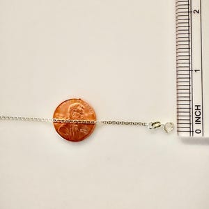 May include: A thin silver chain necklace featuring small, uniform oval links and a spring ring clasp. The chain is positioned horizontally across a copper penny for scale, with a ruler showing measurements in inches along the right side.