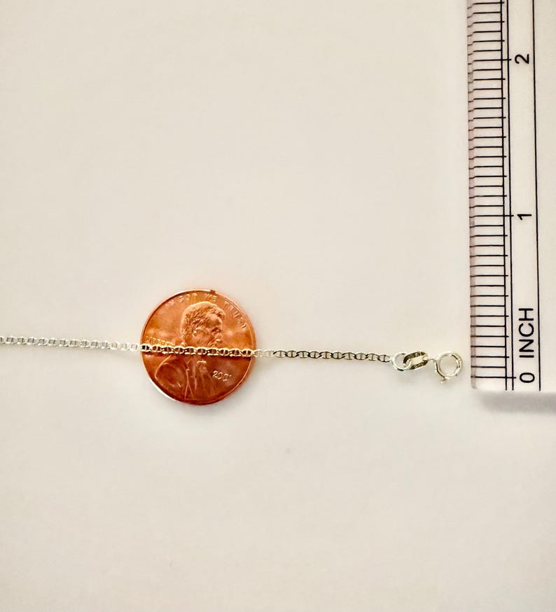 May include: A thin silver chain necklace featuring small, uniform oval links and a spring ring clasp. The chain is positioned horizontally across a copper penny for scale, with a ruler showing measurements in inches along the right side.