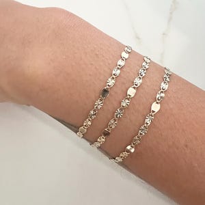 May include: Three delicate gold bracelets with a repeating pattern of small, flat, and star-shaped links. The bracelets are worn on a wrist, showcasing their subtle shine and intricate design. The bracelets are a simple yet elegant accessory.