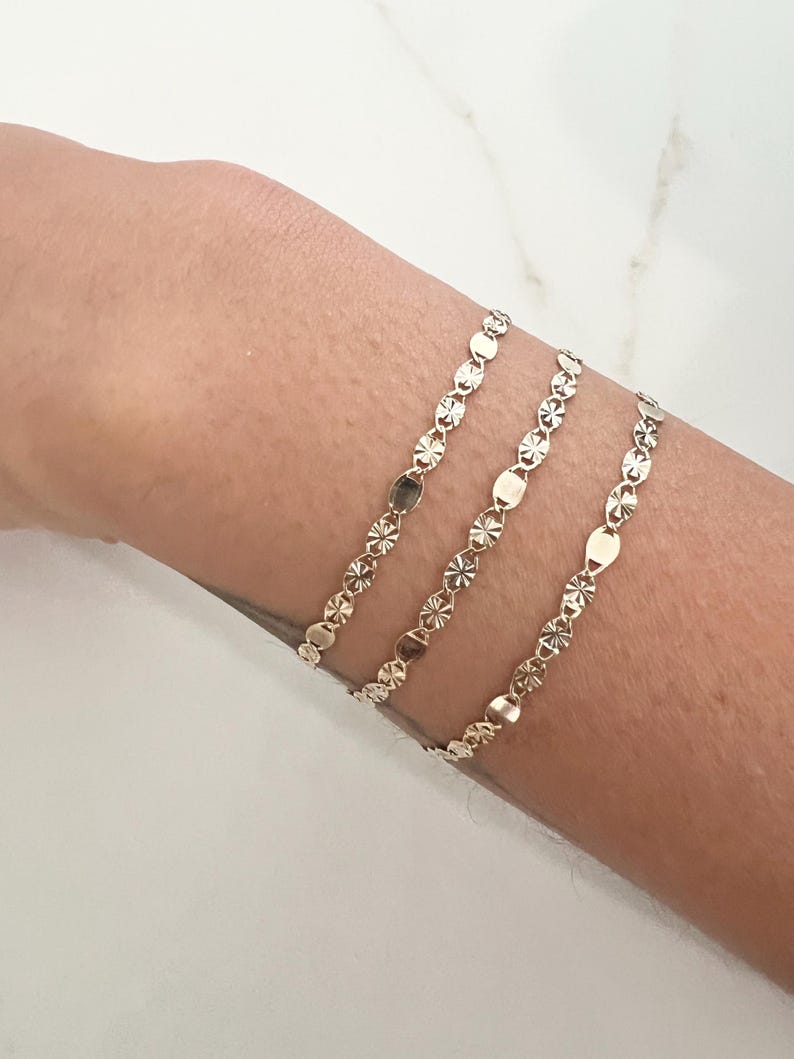 May include: Three delicate gold bracelets with a repeating pattern of small, flat, and star-shaped links. The bracelets are worn on a wrist, showcasing their subtle shine and intricate design. The bracelets are a simple yet elegant accessory.
