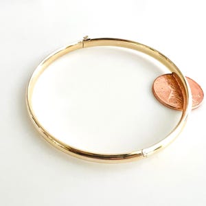 May include: A gold-colored bangle bracelet with a hinged clasp. The bracelet is circular and has a smooth, polished surface. A penny is placed next to the bracelet for size comparison.
