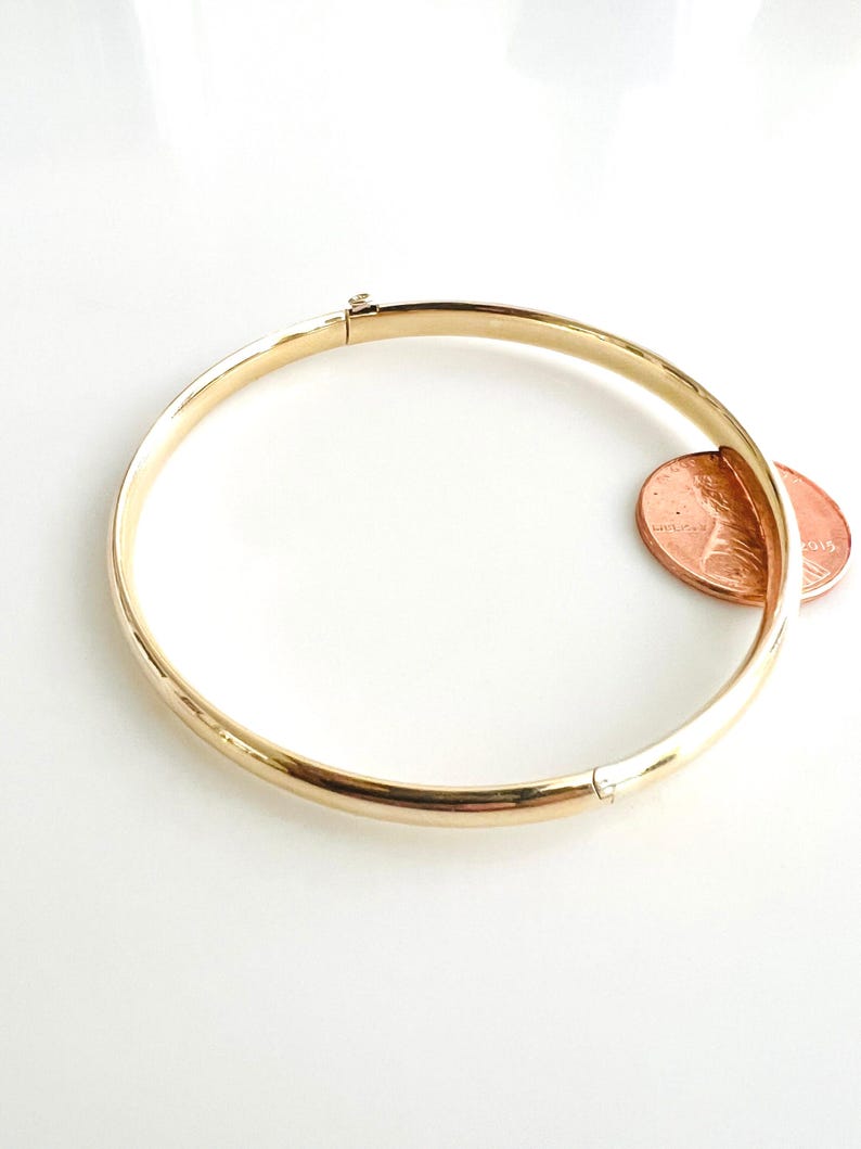 May include: A gold-colored bangle bracelet with a hinged clasp. The bracelet is circular and has a smooth, polished surface. A penny is placed next to the bracelet for size comparison.