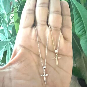Puede incluir: Dos delicados collares con cruces. Uno es plateado, el otro dorado. Cada colgante de cruz cuelga de una fina cadena. Los collares se muestran sobre un fondo de follaje verde.