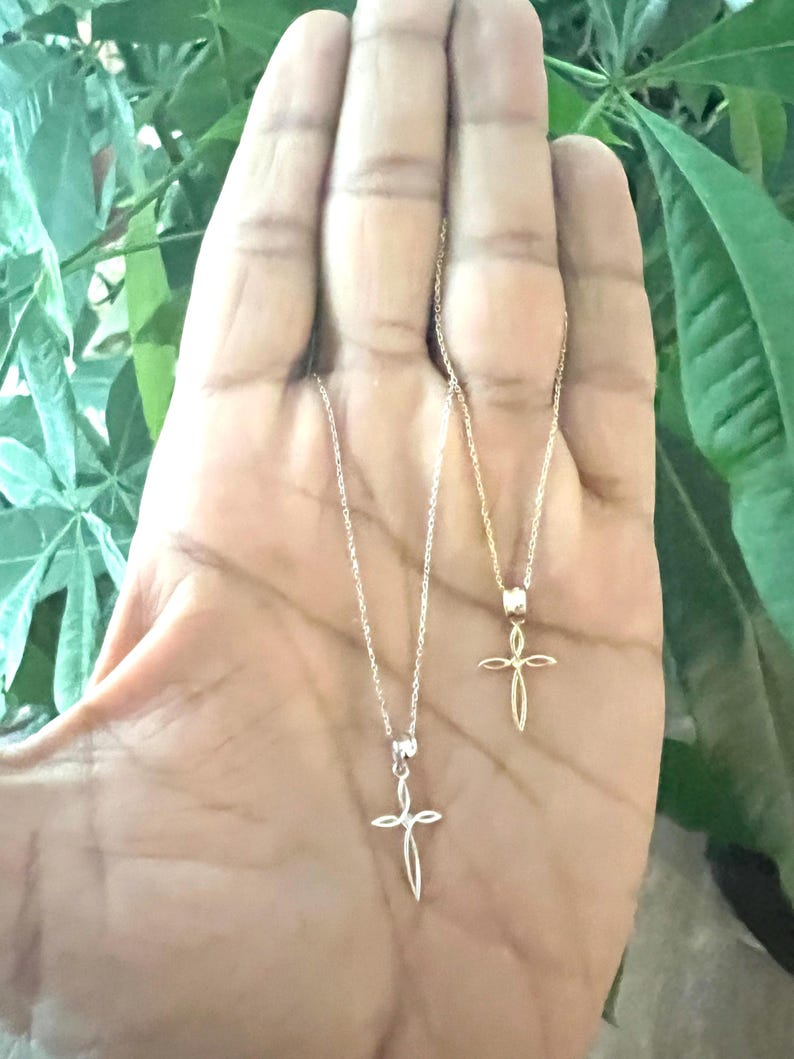 Puede incluir: Dos delicados collares con cruces. Uno es plateado, el otro dorado. Cada colgante de cruz cuelga de una fina cadena. Los collares se muestran sobre un fondo de follaje verde.