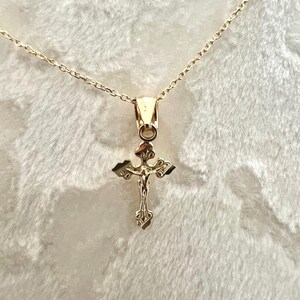 May include: A gold pendant necklace with a detailed crucifix design. The cross features an ornate, textured appearance with a figure of Jesus Christ. The chain is delicate and gold-coloured, creating a cohesive and elegant piece of jewellery.