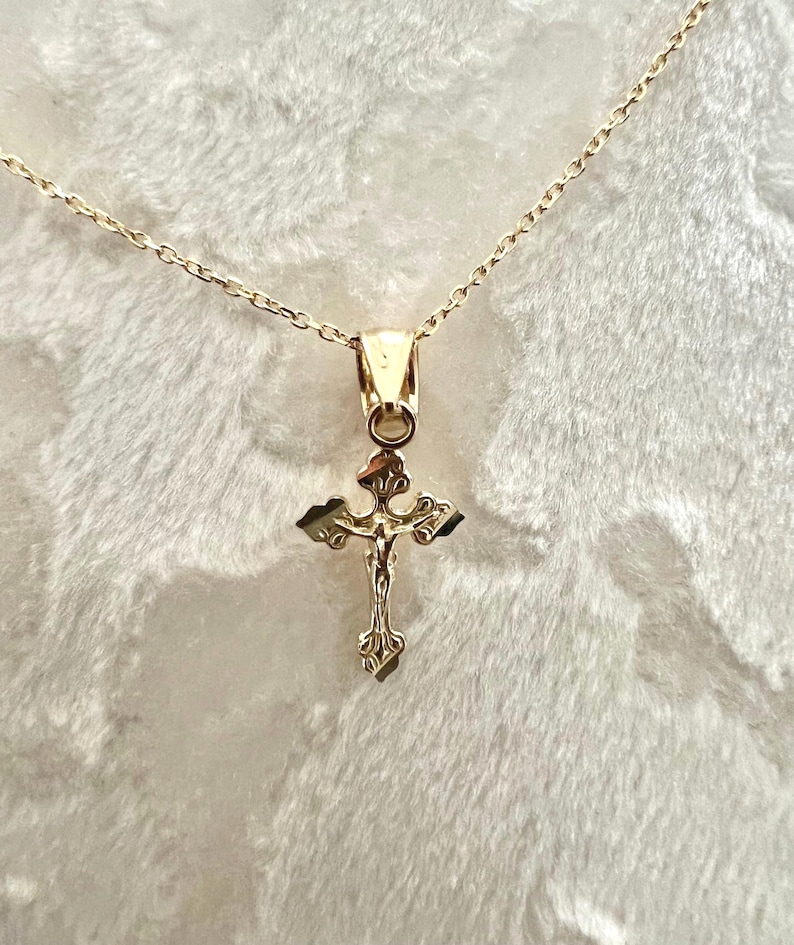 May include: A gold pendant necklace with a detailed crucifix design. The cross features an ornate, textured appearance with a figure of Jesus Christ. The chain is delicate and gold-coloured, creating a cohesive and elegant piece of jewellery.