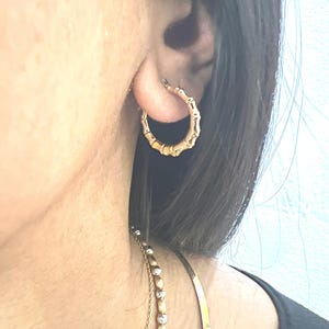Puede incluir: Pendiente de aro dorado con forma de bamb&uacute;. El pendiente se lleva en la oreja y tiene un dise&ntilde;o texturizado que imita el aspecto de los tallos de bamb&uacute;. Tambi&eacute;n se ve un collar de oro con una cadena de cuentas.