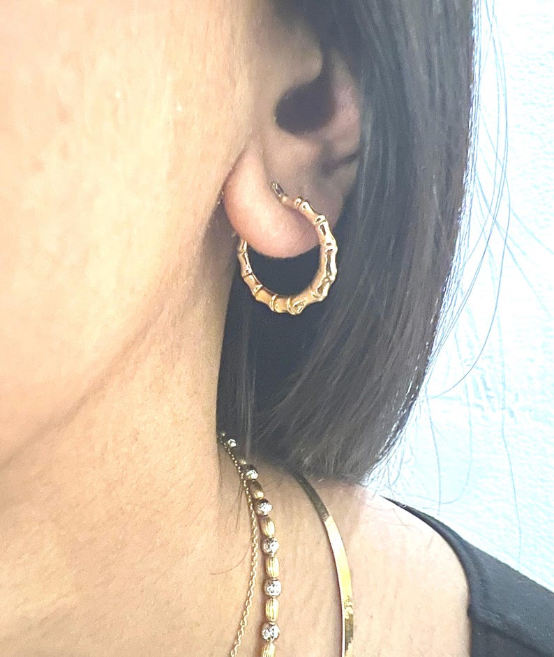 Puede incluir: Pendiente de aro dorado con forma de bamb&uacute;. El pendiente se lleva en la oreja y tiene un dise&ntilde;o texturizado que imita el aspecto de los tallos de bamb&uacute;. Tambi&eacute;n se ve un collar de oro con una cadena de cuentas.