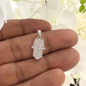 May include: A silver-colored Hamsa hand pendant, encrusted with small, clear gemstones. The pendant is held in a hand, with white flowers in the background. The Hamsa hand is a symbol of protection and good fortune.