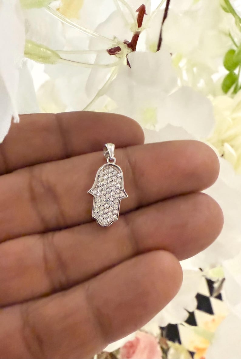 May include: A silver-colored Hamsa hand pendant, encrusted with small, clear gemstones. The pendant is held in a hand, with white flowers in the background. The Hamsa hand is a symbol of protection and good fortune.
