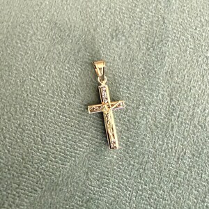 Op de afbeelding: Een goudkleurige kruisvormige hanger met een kleine lus voor een ketting. Het kruis heeft een gedetailleerde afbeelding van Jezus Christus en is versierd met kleine, heldere edelstenen. De hanger staat tegen een gedempte groene achtergrond.