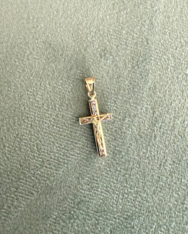 Op de afbeelding: Een goudkleurige kruisvormige hanger met een kleine lus voor een ketting. Het kruis heeft een gedetailleerde afbeelding van Jezus Christus en is versierd met kleine, heldere edelstenen. De hanger staat tegen een gedempte groene achtergrond.