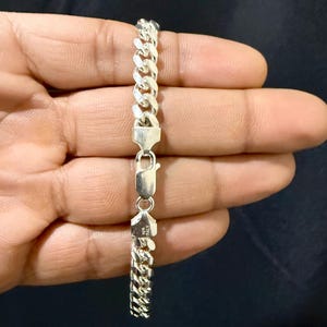 May include: A silver curb chain bracelet with a secure clasp. The bracelet is made of interlocking links and is held against a dark background. The clasp is marked with "925 ITALY".