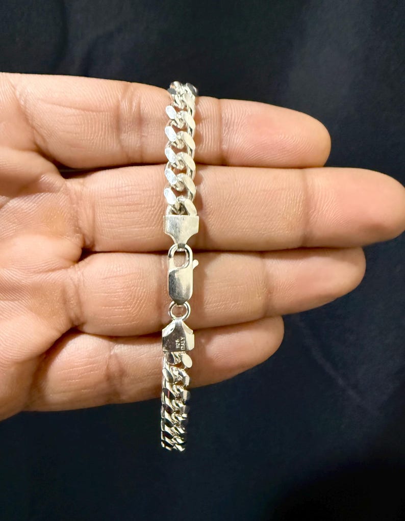 May include: A silver curb chain bracelet with a secure clasp. The bracelet is made of interlocking links and is held against a dark background. The clasp is marked with "925 ITALY".