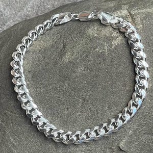 May include: A silver curb chain bracelet. The bracelet is made of interlocking links and has a secure clasp. The jewelry is displayed on a gray, textured surface.