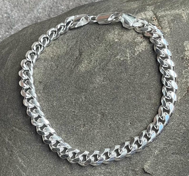 May include: A silver curb chain bracelet. The bracelet is made of interlocking links and has a secure clasp. The jewelry is displayed on a gray, textured surface.